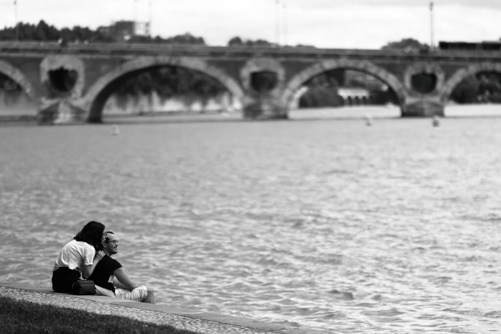 Blog - Les coulisses d'un moment romantique au Port de la Daurade : Un couple en habits élégants, assis par terre, enlacé sur le quai de la Garonne à Toulouse, contemplant la grande roue. Une scène capturée dans l'intimité, révélant l'authenticité de leur complicité.