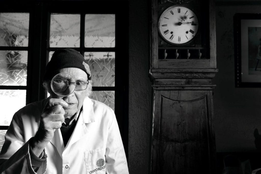 Portrait en noir et blanc de Jacques, 91 ans, chaudronnier et technicien, tenant une loupe devant son œil. L'horloge en arrière-plan et son expression déterminée symbolisent son histoire de résistance et de précision. Projet 'Je suis vieux et je t'emmerde' par Manuel VZ.