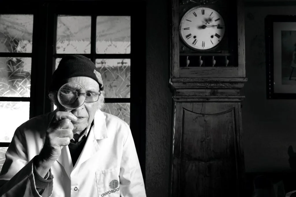 Portrait en noir et blanc de Jacques, 91 ans, chaudronnier et technicien, tenant une loupe devant son œil. L'horloge en arrière-plan et son expression déterminée symbolisent son histoire de résistance et de précision. Projet 'Je suis vieux et je t'emmerde' par Manuel VZ.
