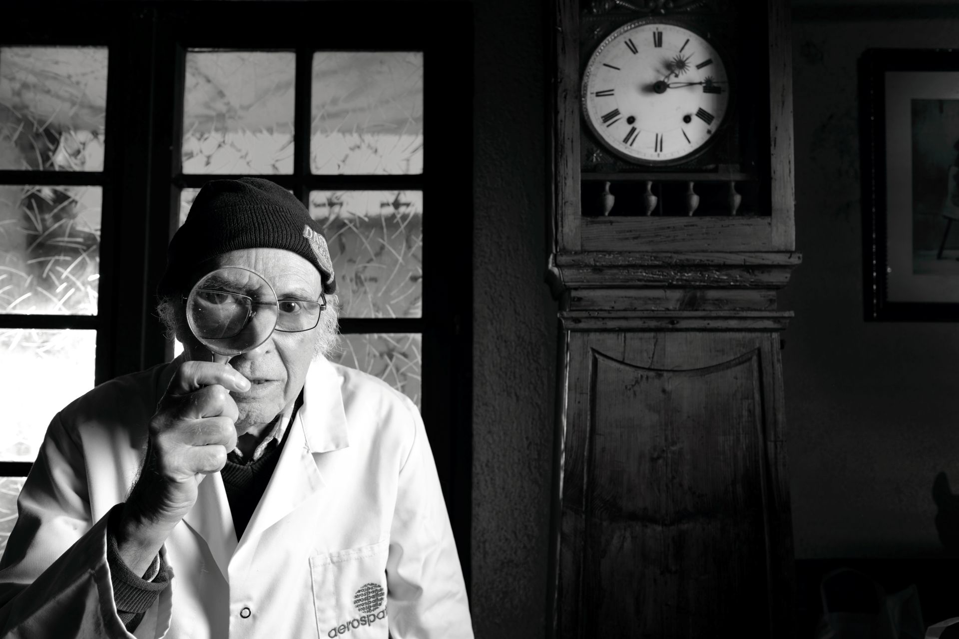 Portrait en noir et blanc de Jacques, 91 ans, chaudronnier et technicien, tenant une loupe devant son œil. L'horloge en arrière-plan et son expression déterminée symbolisent son histoire de résistance et de précision. Projet 'Je suis vieux et je t'emmerde' par Manuel VZ.