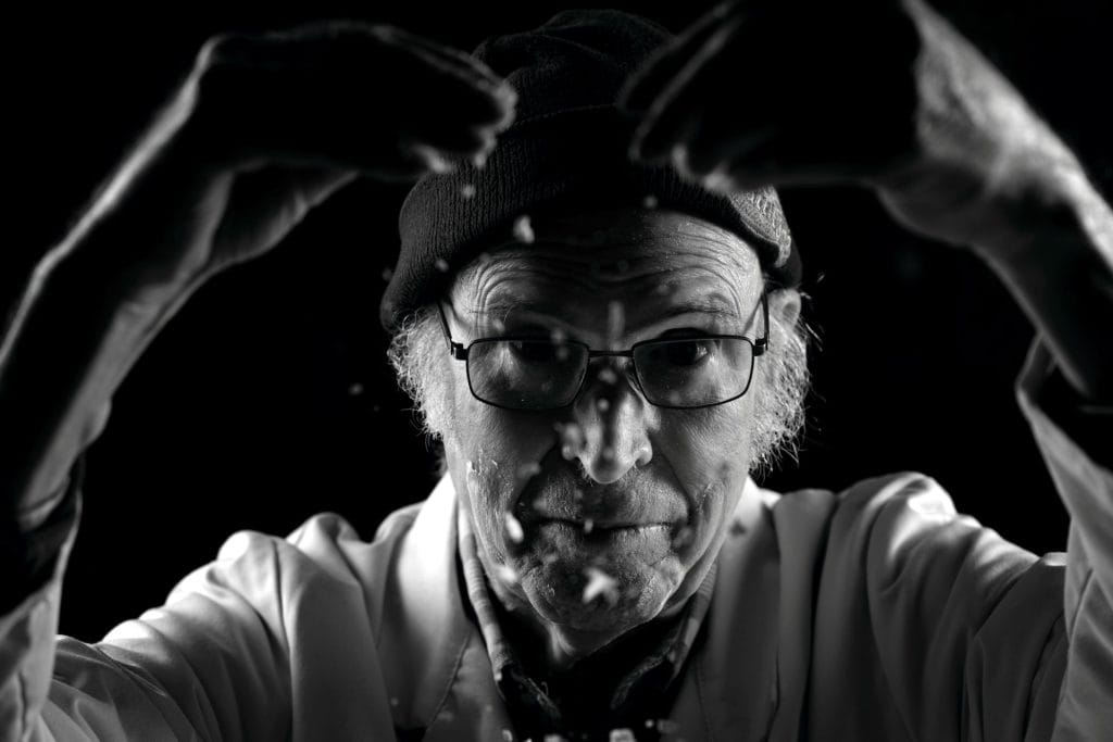 Portrait en noir et blanc de Jacques, 91 ans, les mains levées dans un geste expressif. Les traces de farine ou de poussière autour de lui évoquent son histoire de travail et de résistance. Projet 'Je suis vieux et je t'emmerde' par Manuel VZ.