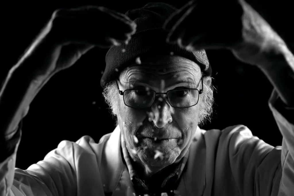Portrait en noir et blanc de Jacques, 91 ans, les mains levées dans un geste expressif. Les traces de farine ou de poussière autour de lui évoquent son histoire de travail et de résistance. Projet 'Je suis vieux et je t'emmerde' par Manuel VZ.
