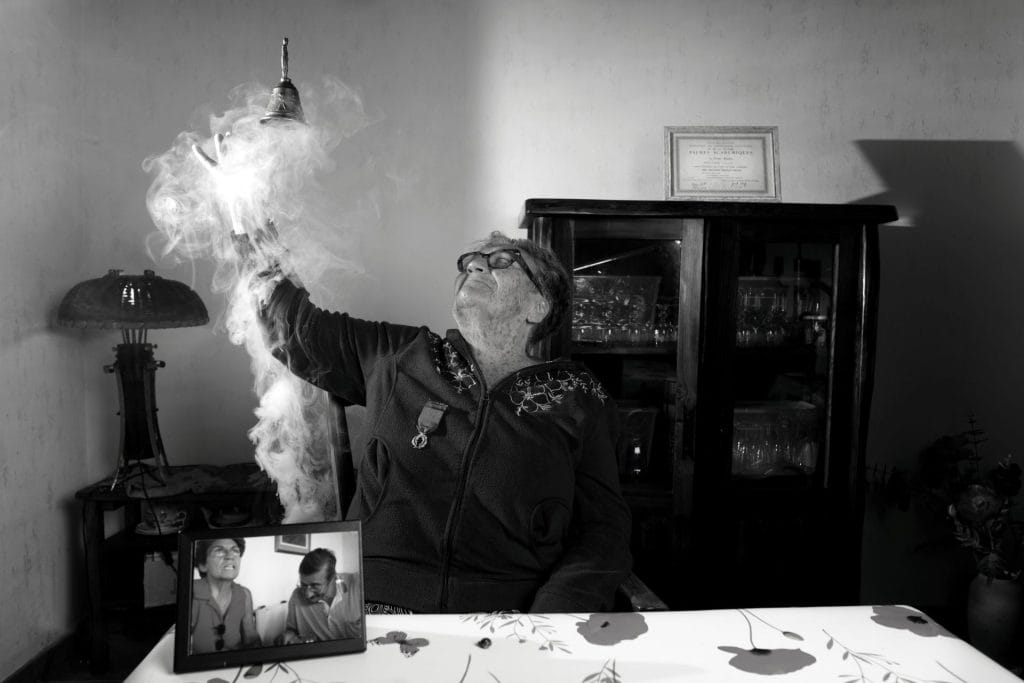 Portrait en noir et blanc d’Hélène, 81 ans, enseignante et militante, tenant une cloche et un cadre avec ses Palmes académiques. Son regard déterminé et son environnement personnel illustrent son engagement pour l’éducation et la justice sociale. Projet 'Je suis vieux et je t'emmerde' par Manuel VZ.
