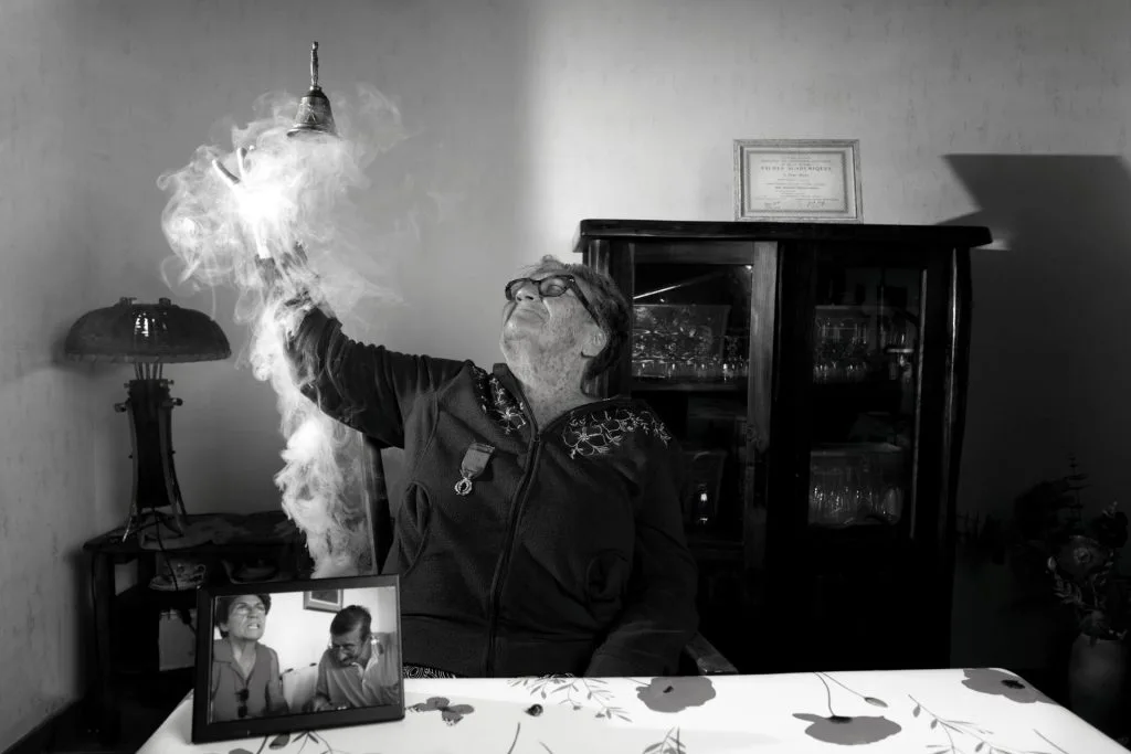 Portrait en noir et blanc d’Hélène, 81 ans, enseignante et militante, tenant une cloche et un cadre avec ses Palmes académiques. Son regard déterminé et son environnement personnel illustrent son engagement pour l’éducation et la justice sociale. Projet 'Je suis vieux et je t'emmerde' par Manuel VZ.