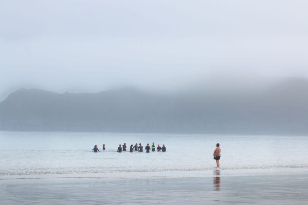 Un groupe de baigneurs immergés dans une mer brumeuse, face à des montagnes estompées. Une scène de communauté et de connexion avec la nature, capturant l’émotion des rivages bretons ou basques.