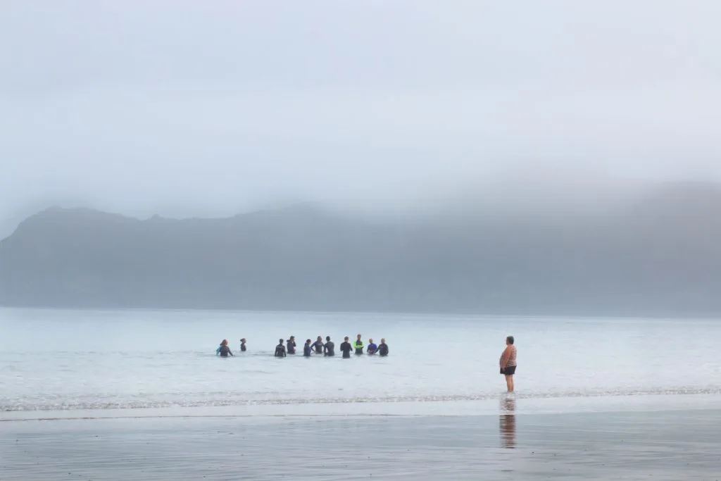 Un groupe de baigneurs immergés dans une mer brumeuse, face à des montagnes estompées. Une scène de communauté et de connexion avec la nature, capturant l’émotion des rivages bretons ou basques.