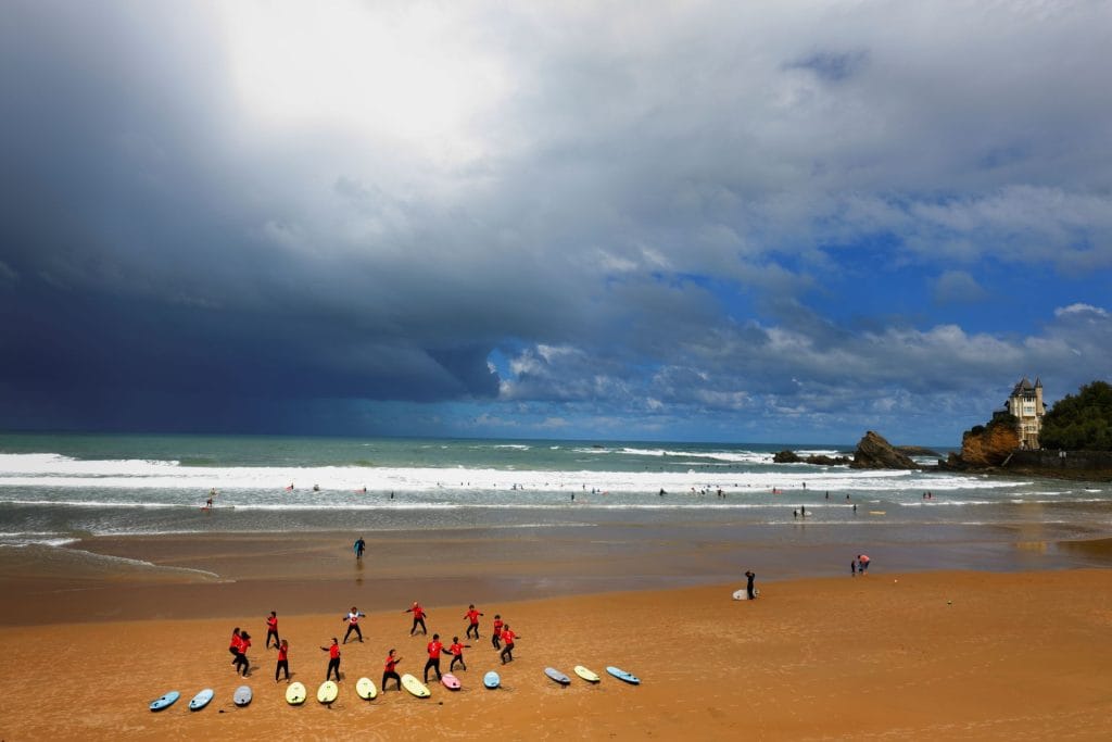 Une plage animée de Biarritz sous un ciel orageux. Des surfeurs en combinaison rouge préparent leurs planches, tandis que les vagues déferlent sur le sable doré. Une scène dynamique du Pays Basque, où l’énergie de la mer rencontre celle des hommes.