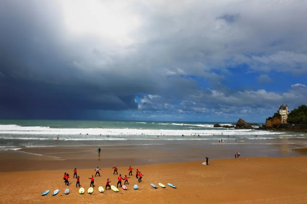 Une plage animée de Biarritz sous un ciel orageux. Des surfeurs en combinaison rouge préparent leurs planches, tandis que les vagues déferlent sur le sable doré. Une scène dynamique du Pays Basque, où l’énergie de la mer rencontre celle des hommes.